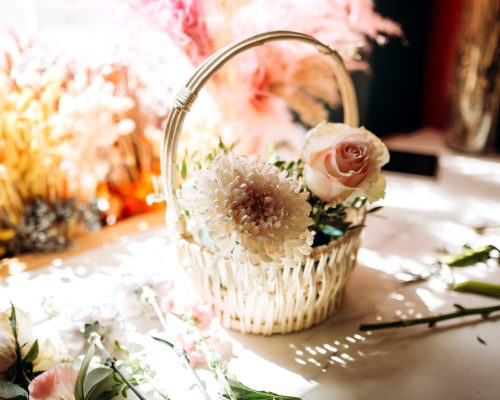 There is a little white basket with white peony and rose on the wooden table in the flower shop .